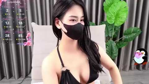 Media: A video of an East Asian woman with long black hair, wearing a black mask and revealing black bikini top, sitting on a beige couch. Background features a green plant and grey curtains.