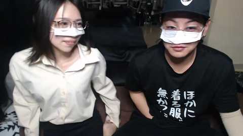 Media: Video of two women wearing surgical masks and white lab coats, sitting side by side in a dimly lit room with black furniture.