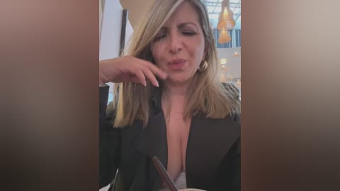 Media: Video of a blonde woman with medium build, wearing a black blazer and a low-cut shirt, making a face while holding a phone, in a dimly lit restaurant with warm lighting and hanging lamps.