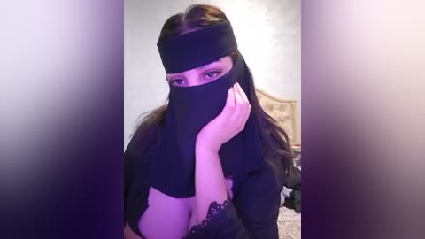 Media: Video of a woman with long, dark hair, wearing a black hijab and headband, covering her face partially. She has light skin and is indoors, possibly in a bedroom with a beige headboard visible in the background.