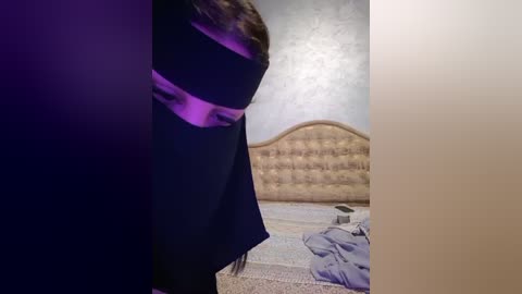 Media: Video of a woman wearing a black dress, with a purple glow on her face, standing near a beige tufted headboard in a dimly lit room with scattered clothes on the floor.