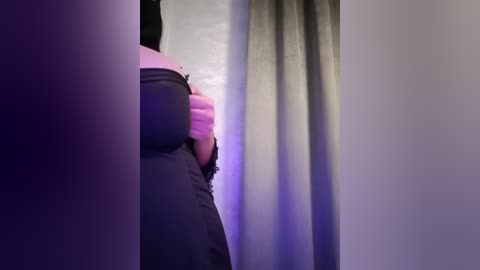 Media: A video showing a person's head and upper torso partially obscured by a dark curtain. The person, wearing a pink shirt, is leaning against a wall with a light purple hue. The scene is dimly lit, creating a moody atmosphere.