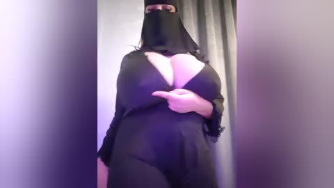 Media: Video of a plus-size woman with a pale complexion wearing a sheer black burqa, her large breasts partially exposed, standing in a dimly lit room with purple curtains.