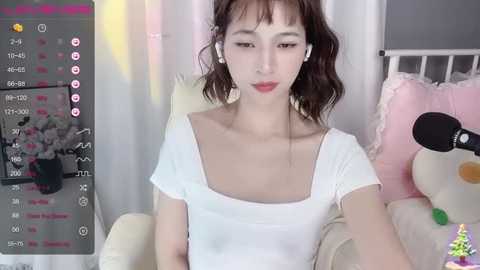 Media: Video of a young Asian woman with fair skin, short wavy brown hair, and a small bust, wearing a white short-sleeved top. She sits on a pastel-colored couch, with a plush Minnie Mouse toy beside her. A smartphone screen displaying a live stream is visible in the background.