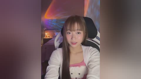 Media: Video of an East Asian woman with long, straight brown hair, wearing a white cardigan over a pink tank top, sitting in a gaming chair, in a dimly lit room with colorful LED lights.