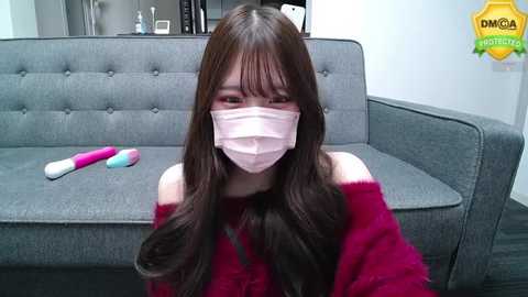 Media: Video of an Asian woman with long brown hair, wearing a pink mask, red sweater, and seated on a gray sofa. Pink vibrator and toys on the couch.