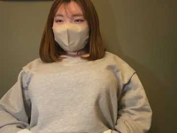 Media: Video of a young woman with straight, shoulder-length brown hair, wearing a light gray sweater and a face mask, standing against a muted green wall.