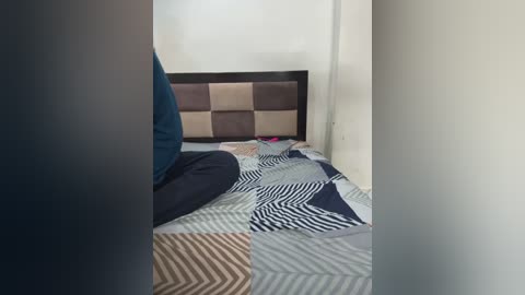 Media: Video of a person sitting on a bed with a patterned quilt, wearing a blue shirt and dark pants, against a plain white wall.