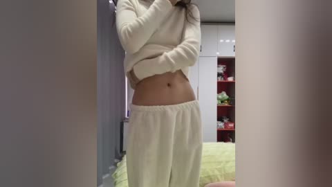 Media: Video of a young woman with light skin, medium build, wearing white fleece pajama pants and a matching top, standing in a bedroom with white cabinets and green bedding.