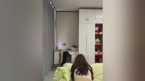 Video of a modern bedroom with a white desk, black chair, white cabinets, red shelves, green bedspread, and a woman with long dark hair in a white top.