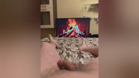 Media: Video of a nude man lying on a bed with a patterned comforter, legs spread, and a TV displaying a fire background in the background.