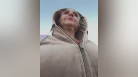 Media: A video of a middle-aged woman with fair skin and short blonde hair, wearing a beige hooded jacket with a red zipper, standing outdoors against a clear blue sky.