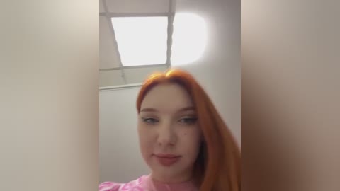 Media: Video of a young Caucasian woman with long, straight, bright red hair, wearing a pink top, standing in a brightly lit hallway with fluorescent lighting.