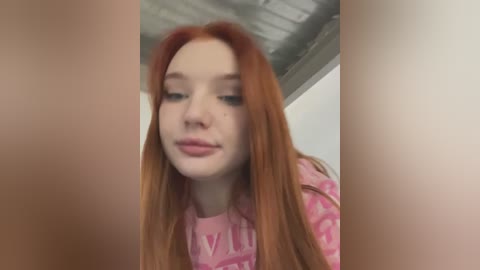 Media: A close-up video of a young woman with long, straight, red hair, fair skin, and light makeup, wearing a pink shirt with white text. The background features a ceiling with exposed beams.