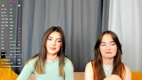 Media: Video of two young women with light skin, straight brown hair, and wearing light blue long-sleeve tops, seated against a grey curtain backdrop.