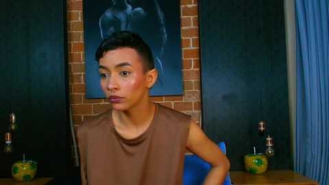 Media: Video of a young Asian man with short black hair, wearing a brown sleeveless shirt, seated in front of a brick wall with a large black and white painting and two green ceramic vases on a wooden table.