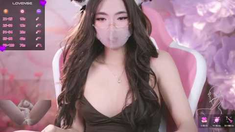 Media: A video of an Asian woman with long, wavy black hair, wearing a black dress, pink cat ears, and a mask, sitting in a pink gaming chair. Background features soft pink and white hues.