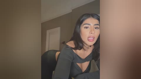 Media: Video of a young Latina woman with long black hair, wearing a black off-shoulder crop top, leaning provocatively against a door frame in a dimly lit room.