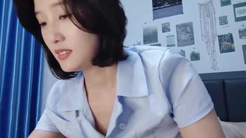 Media: Video of a young East Asian woman with shoulder-length black hair, wearing a light blue blouse, in a bedroom with blue curtains and anatomical posters on the wall.