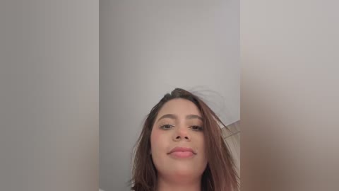 Video of a young Latina woman with light brown skin, straight brown hair, and light makeup, smiling. She stands in a minimalist, white-walled room with a blurred, beige door in the background.