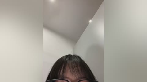 Media: Video of an Asian woman with straight black hair and glasses, partially visible from the neck up, in a minimalist, white-walled room with recessed ceiling lights.