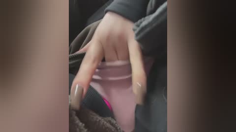 Media: Video of a person's hands holding a pink iPhone, with a blurry background featuring a dark jacket and a textured, gray blanket.