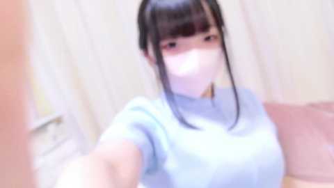 A blurry video of an East Asian woman with straight black hair, wearing a light blue top and face mask, taking a selfie indoors. The background is indistinct.