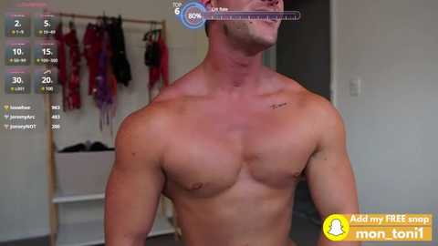 Media: Video of a shirtless, muscular white man with a small tattoo on his shoulder, standing indoors. Background includes a closet with hanging clothes, a laundry basket, and a digital fitness app overlay displaying workout metrics.