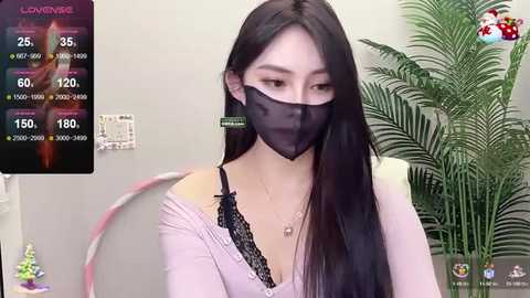 Media: Video of an East Asian woman with long black hair, wearing a black face mask, a light pink top, and black lace bralette, seated in a modern room with a green plant in the background.