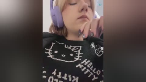 Media: Video of a young woman with fair skin, wearing a black sweater with white \"Kitty\" logo, purple headphones, and a contemplative expression.