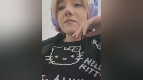 Media: A video of a young woman with short, dyed pink hair, wearing a black sweater with a white Hello Kitty design. Her face is neutral, and she has a hand on her neck.