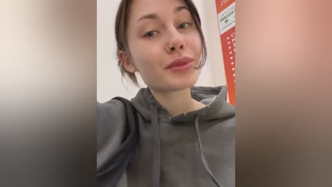 Media: Video of a young woman with fair skin, dark hair, and a nose ring, wearing a gray hoodie, taken from a selfie angle. Background shows a blurred, red and white sign.