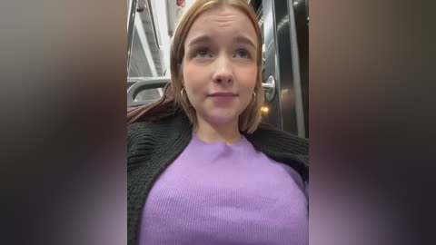 Media: Video of a young Caucasian woman with light skin and straight, blonde hair, wearing a purple knit sweater and black cardigan, sitting in a subway car.