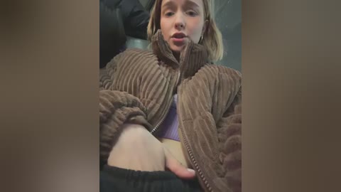 Media: A video shows a young Caucasian woman with fair skin, blonde hair, and a neutral expression. She's wearing a brown quilted jacket and black pants, with her legs spread. The background is blurred, focusing on her.