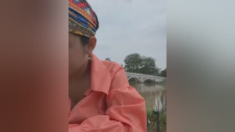 Media: Video of a person in a bright coral shirt, wearing a colorful, patterned headdress, standing by a serene lake with a stone bridge in the background. The image is slightly blurred, focusing on the person's profile and the peaceful scenery.