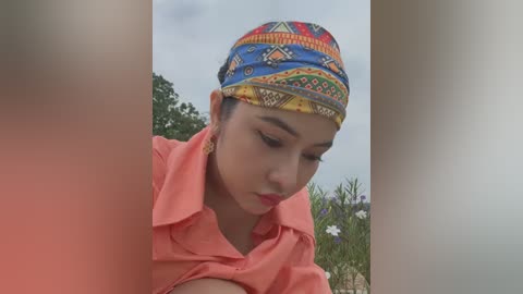 Media: Video of a young woman with light brown skin, wearing a colorful, patterned headscarf and a coral shirt, focusing intently. Background features blurred foliage and a cloudy sky.