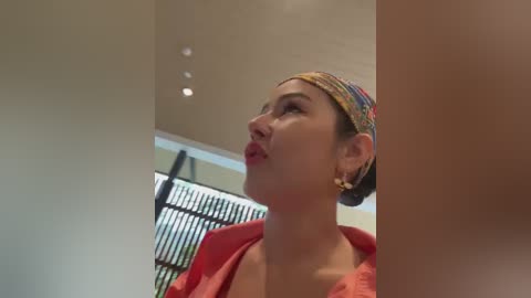 Media: Video of a woman with light skin, dark hair tied back, wearing a red blouse and colorful headscarf, looking upward in a modern, minimalist room with a beige ceiling and large windows.