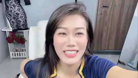 Media: A video of a smiling Asian woman with straight black hair, wearing a navy blue shirt with yellow trim, taking a selfie indoors, featuring a white chair, wooden door, and floral decorations.