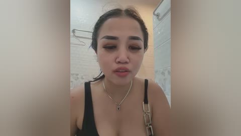 Media: Video of an East Asian woman with light skin, black hair tied back, wearing a black tank top and a silver necklace, standing in a tiled bathroom with white walls and a metal towel rack.