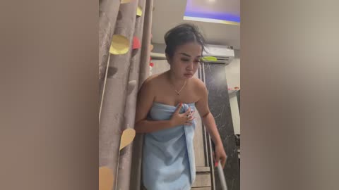 Media: Video of a young Asian woman with dark hair in a bun, wearing a light blue towel, standing in a bathroom with beige curtains and a colorful shower curtain.