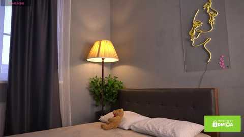 Media: Video of a cozy bedroom with a tufted brown headboard, a small teddy bear, and a potted plant. A yellow lampshade casts a warm glow on the gray walls adorned with a minimalist abstract art piece.