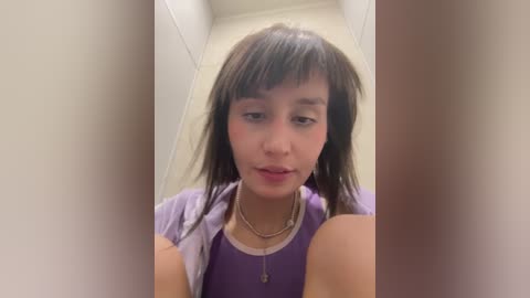 Media: Video of a young, light-skinned woman with straight, dark hair and bangs, wearing a purple top and gold necklace, standing in a narrow, tiled hallway.