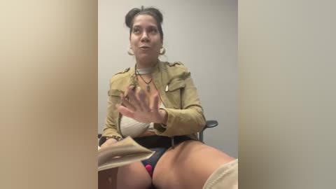 Media: Video of a woman in a beige jacket, black shorts, and beige high-heeled shoes, sitting on a black chair, gesturing with her hands, in a plain white room.