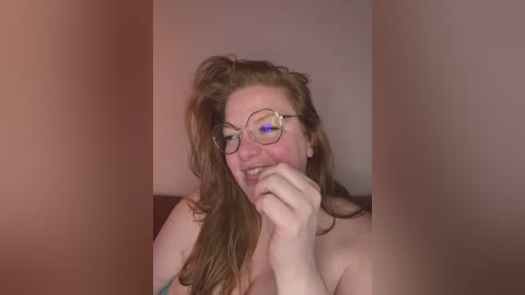 Media: Video of a fair-skinned, red-haired woman with glasses, wearing a pink top, playfully covering her mouth with her hand. The background is blurred with warm lighting, creating a cozy indoor setting.