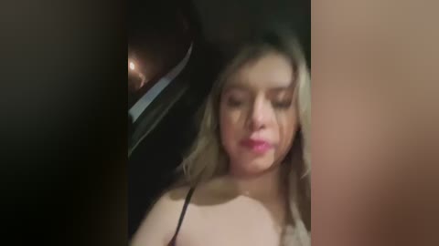 Media: A blurry video of a young woman with light skin and shoulder-length blonde hair, wearing a black spaghetti-strap top, seated in a dimly lit car.