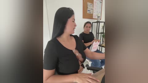 Media: Video of two women in a classroom: one with long black hair, wearing a black top, holding a clipboard, and another with glasses, sitting at a desk, holding a pink folder.