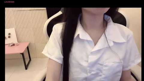 Media: A video of an East Asian woman with long black hair, wearing a white short-sleeved blouse, seated in a gaming chair with a pink desk and beige tiled wall in the background.