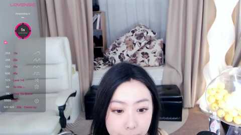 Media: Video of an East Asian woman with long black hair, makeup, and a beige top, sitting in a living room with floral pillows, white chair, and a bowl of yellow fruit.