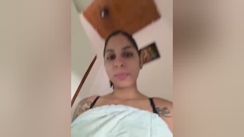 Media: Video of a young, light-skinned woman with dark hair, wearing a light blue towel, showcasing tattoos on her arms, standing in a room with a wooden ceiling fan and framed pictures.