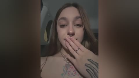 Media: Video of a young woman with long brown hair, light skin, and a tattoo on her chest, covering her mouth with her hand in a dimly lit car interior.